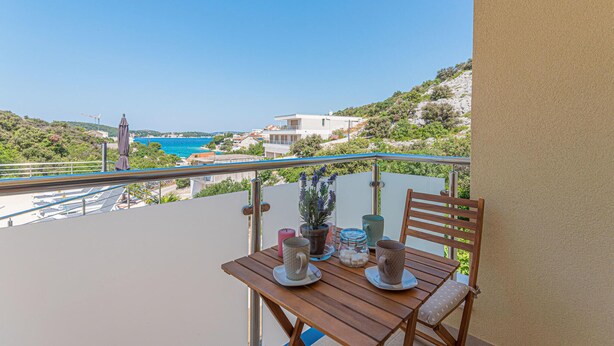 Un balcon avec une table en bois, dressée pour deux personnes, offrant une vue sur la côte, avec des bâtiments et de la verdure.