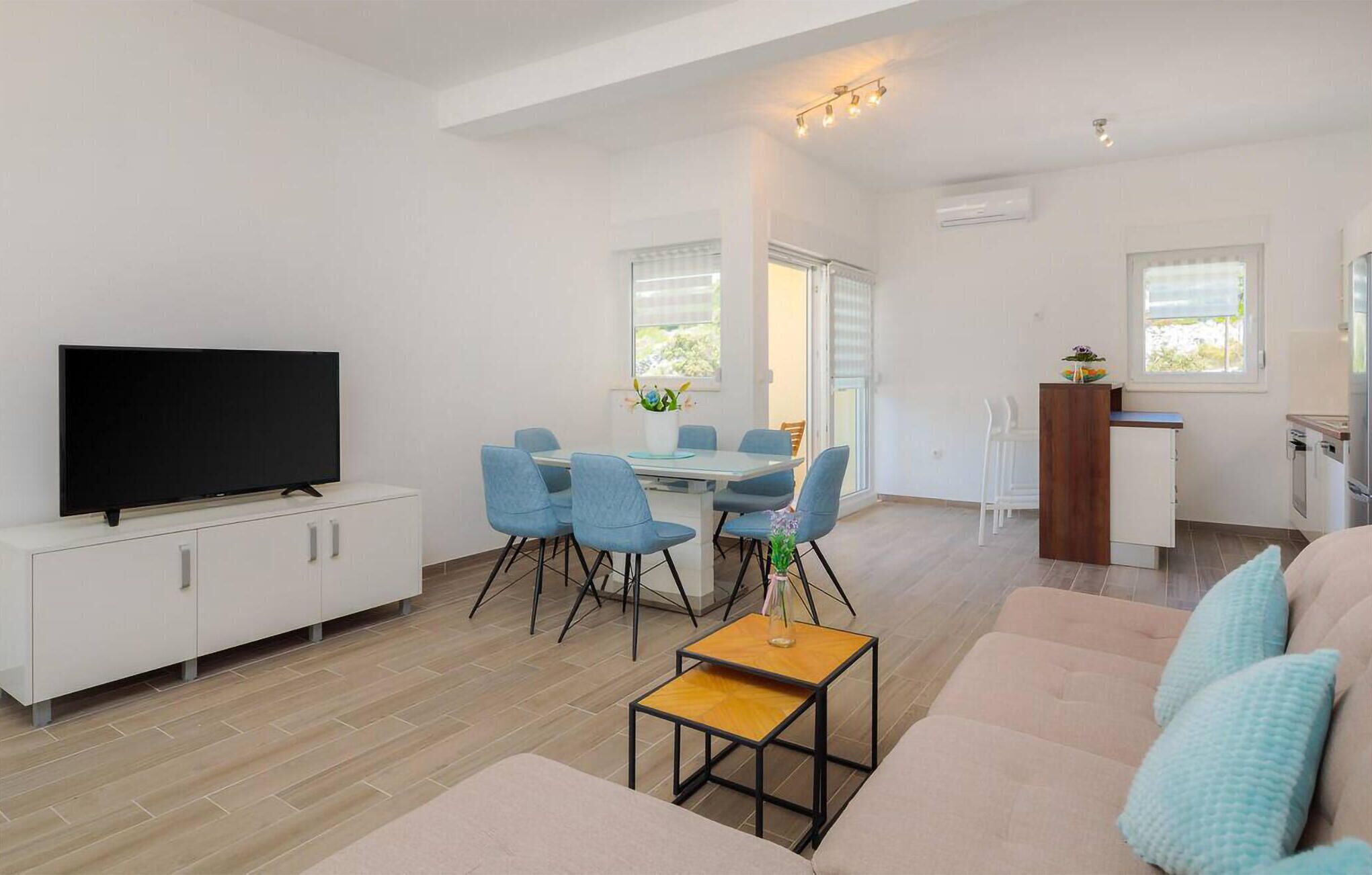 Een moderne woonkamer met een flatscreen televisie, een eethoek met blauwe stoelen, een houten salontafel en een kitchenette met een houten aanrecht.