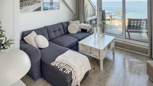 Een moderne woonkamer met een grijze bank, witte kussens, een witte salontafel en uitzicht op zee door grote ramen.