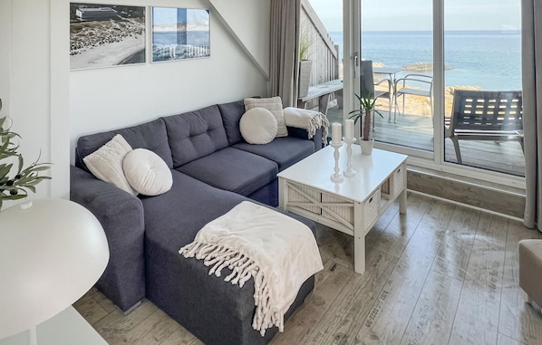 Un salon moderne avec un canapé gris, des coussins blancs, une table basse blanche et une vue sur la mer grâce à de grandes fenêtres.