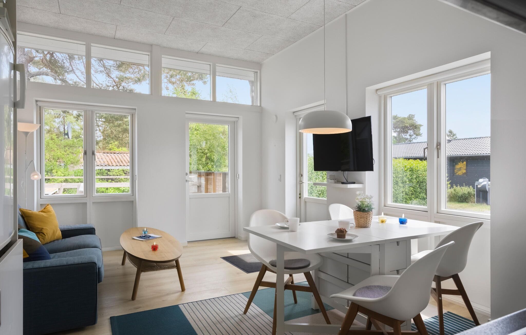 Ein heller, moderner Wohnraum mit einer Couch, einem Couchtisch und einem Esstisch mit Stühlen. Große Fenster bieten natürliches Licht und Blick ins Grüne.