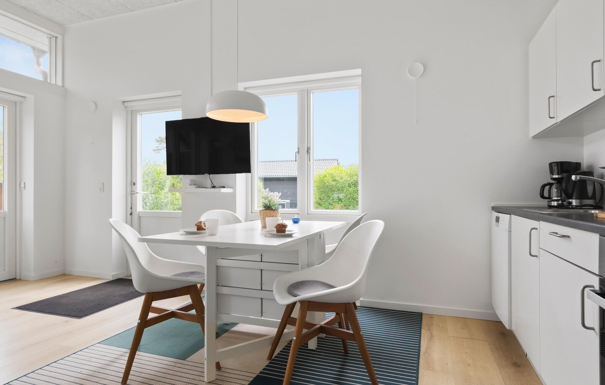 A modern kitchen with white cabinets, a dining table with two chairs, a black TV on the wall, and a coffee maker on the counter.
