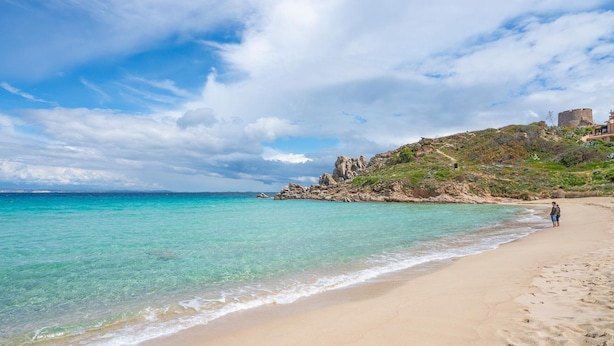 Una playa con agua turquesa cristalina, una orilla arenosa y terreno rocoso con edificios en una colina.