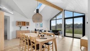 Een ruime eethoek met een houten tafel en stoelen, een hanglamp en uitzicht op het buitenlandschap door grote schuifdeuren.