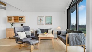 Een moderne woonkamer met een zithoek, een houten salontafel en uitzicht op de omgeving door grote ramen.