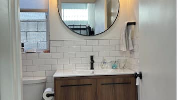 Features a sleek, white-tiled bathroom with a wooden vanity and round mirror.