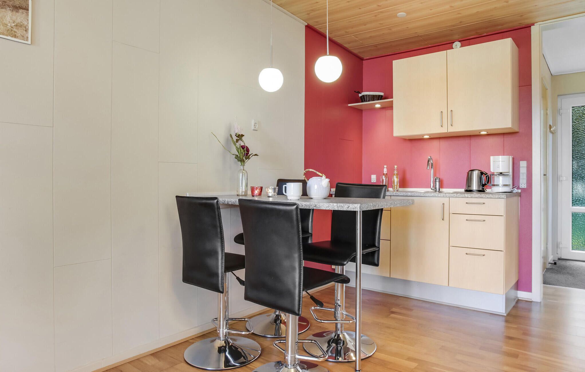 Een moderne keuken met een rode accentmuur, een eethoek met twee zwarte stoelen en een koffieapparaat.