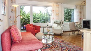 A spacious living room with a red sofa, a white armchair, and a glass coffee table. Large windows offer a view of greenery outside. - Awesome apartment in Goslar with WiFi (Goslar)
