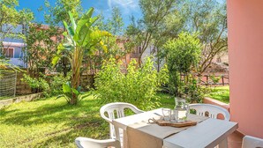 Um pátio externo com mesa e cadeiras, cercado por vegetação exuberante e árvores.