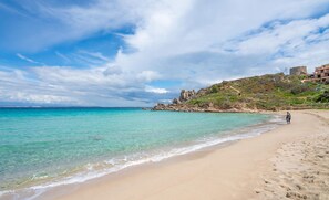Una playa con agua turquesa cristalina, una orilla arenosa y terreno rocoso con edificios en una colina.