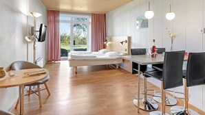 Una habitación de hotel moderna con una cama, una mesa de comedor con sillas, un escritorio con una silla y un ventanal con cortinas.