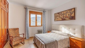 Een slaapkamer met een bed, een stoel, een nachtkastje, een radiator, een raam met gordijnen en een schilderij aan de muur.