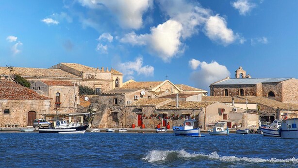 Un pueblo costero con edificios históricos de piedra, un cielo azul despejado y varias embarcaciones amarradas a lo largo del muelle.