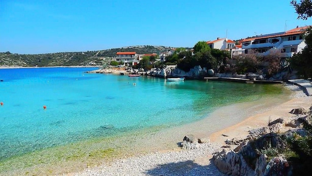 A coastal area with clear turquoise waters, a pebble beach, and buildings with red roofs. - 2 bedroom beach front apartment in Rogoznica (Rogoznica)