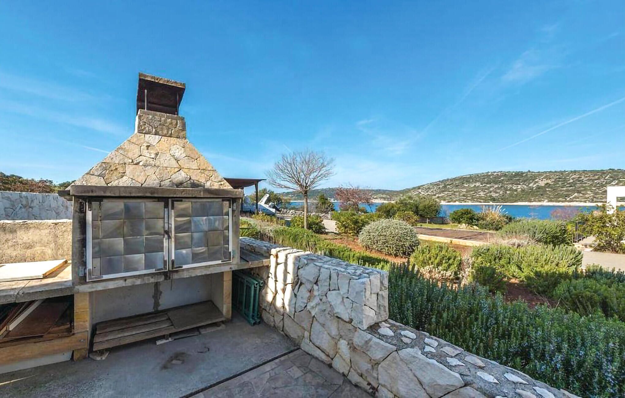 A stone-built outdoor kitchen with a chimney, surrounded by a stone wall and lush greenery.