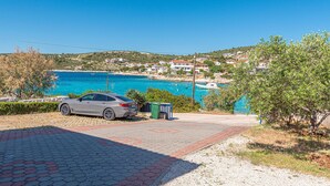 A coastal road with a parked car, a trash bin, and a clear view of a seaside village with houses and a boat in the water. - 2 bedroom beach front apartment in Rogoznica (Rogoznica)