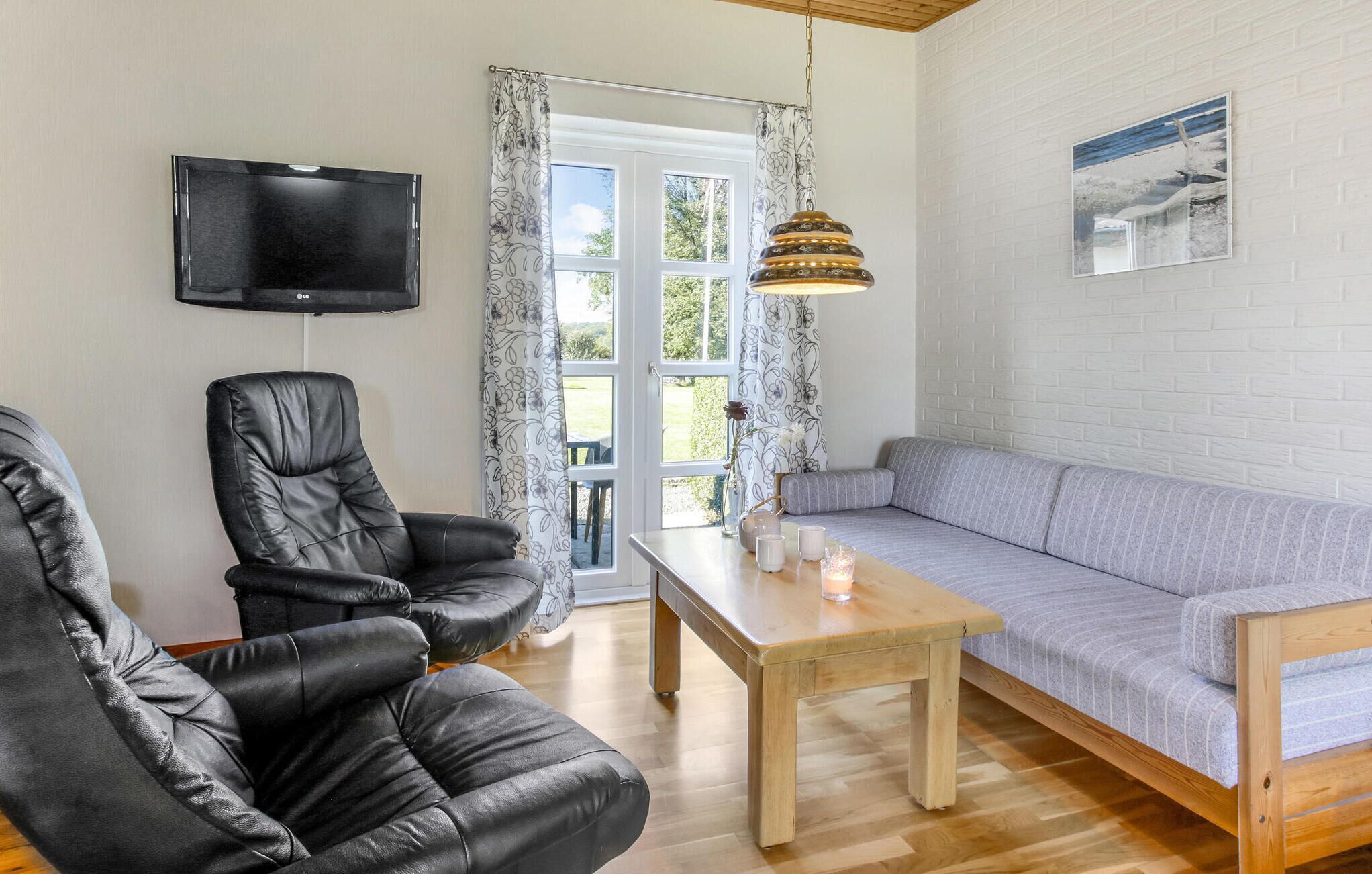 A living room with a wooden floor, a grey sofa, a black leather chair, a wooden coffee table, a TV mounted on the wall, and a window with curtains.