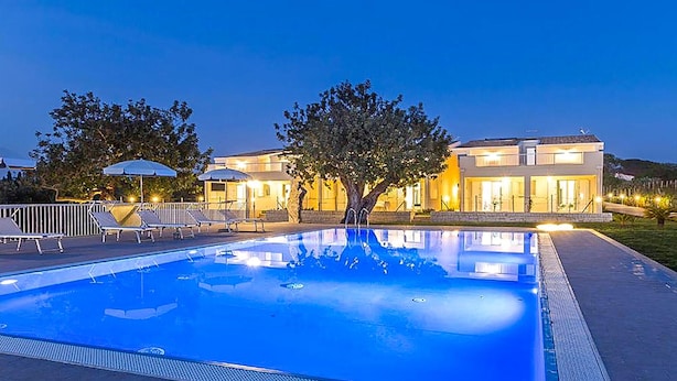 Ein Swimmingpool mit Liegestühlen und Sonnenschirmen, umgeben von einem Gebäude mit mehreren Balkonen und Beleuchtung.