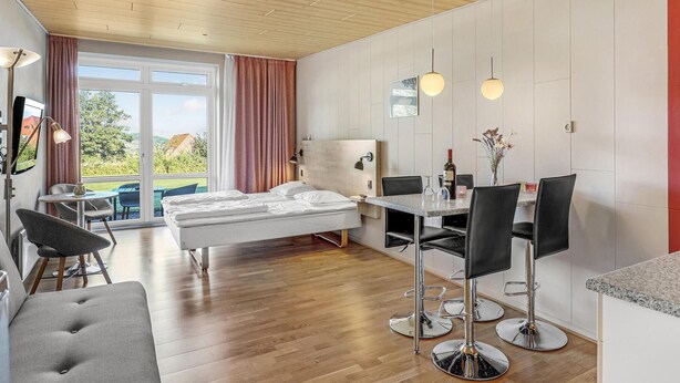 A modern hotel room with a bed, a desk with two chairs, a dining table, and a view of the outdoors.
