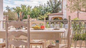 Ein Holztisch mit einem gewobenen Stuhl, eine Fruchtschale und eine Tasse auf einem Balkon mit Blick auf einen Garten und ein Gebäude.