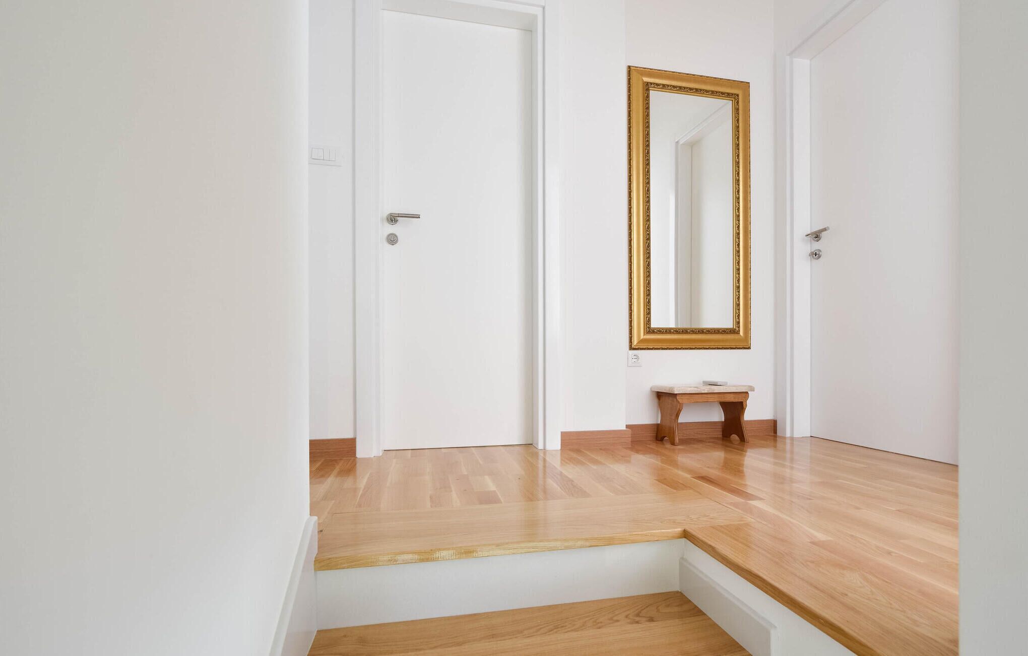 Um corredor com piso de madeira, porta branca e espelho com moldura dourada.