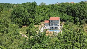 A two-story house with a red roof, surrounded by dense green trees, and a swimming pool in the front yard. - Stunning home in Sinj (Sinj)