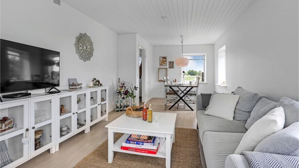A modern living room with a flat-screen TV, white cabinetry, a coffee table with books, a grey sofa, and a dining area with a table and chairs. - 1 bedroom nice home in Rønne (Rønne)