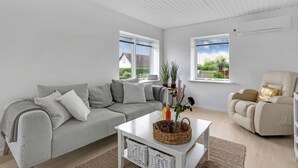 A modern living room with a grey sofa, a white coffee table, and a beige armchair. There are two windows providing views of the outside. - 1 bedroom nice home in Rønne (Rønne)