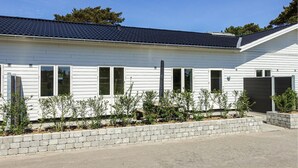 A modern single-story house with a dark roof, white siding, and a garage. There are small shrubs in front of the house. - Amazing apartment in Nexø with kitchen (Nexø)