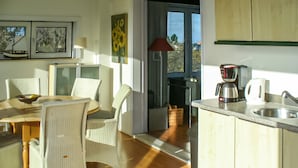 Une cuisine lumineuse équipée d’une machine à café, un coin repas avec des chaises en osier et une vue par la fenêtre.