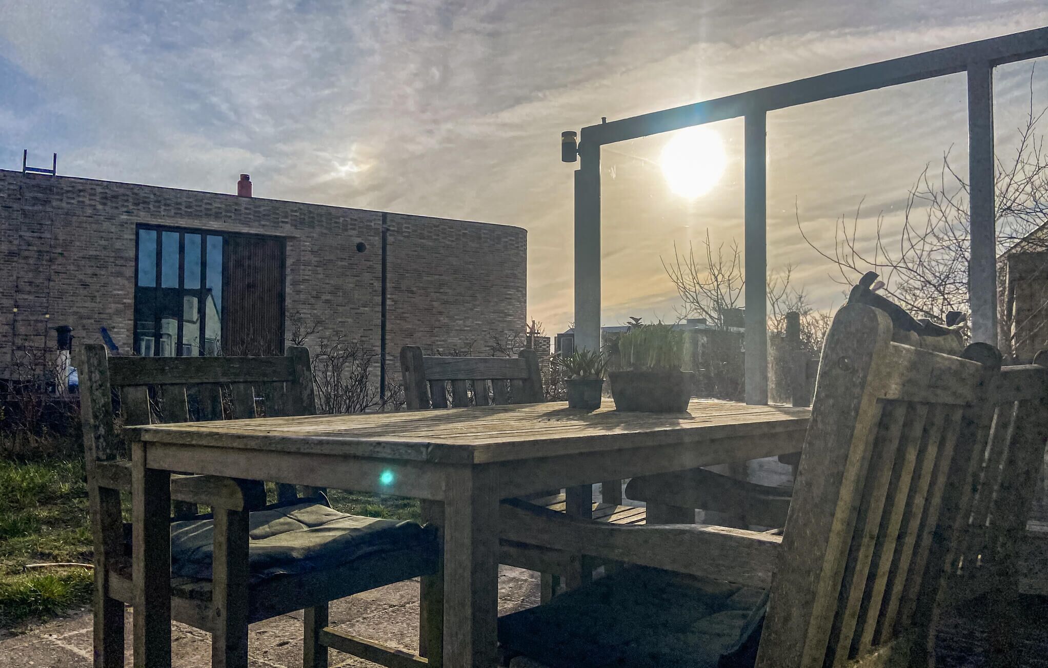 A wooden table and chairs in an outdoor setting with a brick building in the background and the sun shining through a window.