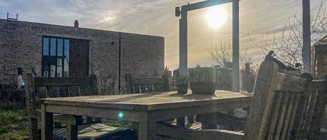 A wooden table and chairs in an outdoor setting with a brick building in the background and the sun shining through a window.