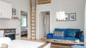 Eine moderne Wohnung mit Küche, Essbereich und Wohnzimmer. Es gibt ein Schlafbett im Hochbettbereich mit einer Leiter, ein blaues Sofa mit gemusterten Kissen und eine Pendelleuchte.