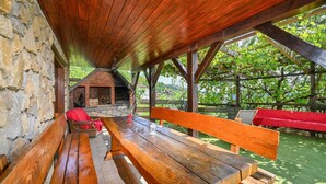 Ein rustikaler Sitzbereich im Freien mit einer Steinmauer, Holzbanken und einem Kamin aus Ziegelsteinen unter einem Holzdach.