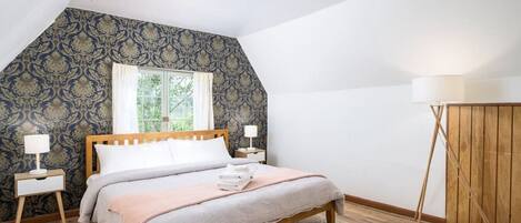 A bedroom with a wooden bed, two bedside tables with lamps, a window with curtains, and a patterned wallpaper.