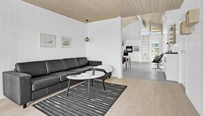 Ett modernt vardagsrum med en svart lädersoffa, ett runt soffbord och en svartvit randig matta. Det finns också en köksdel med vita skåp och ett matbord med stolar.