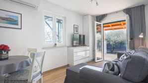 Una sala de estar moderna con un sofá, una mesa de comedor con sillas, un televisor sobre un mueble bajo y un ventanal con cortinas.