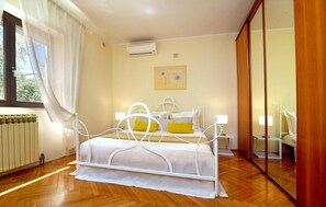 A bedroom with a white bed, a bedside table with a lamp, a radiator, and a window with sheer curtains. - Gorgeous home in Pula (Pula)
