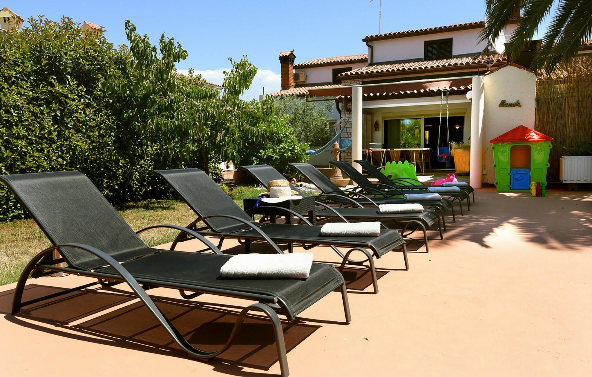 A row of sun loungers with towels, a small playhouse, and a building with a tiled roof.