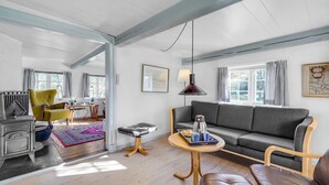 Una sala de estar con sofá gris, mesa de centro de madera y sillones de cuero. Se ve una estufa antigua en la esquina, y en el fondo un área de comedor con una silla y mesa amarillas.