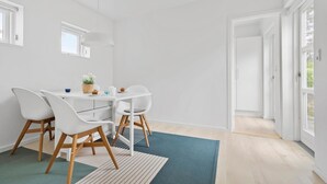 Une pièce lumineuse et blanche, avec une table à manger, des chaises et une vue sur l’extérieur par une porte ouverte.
