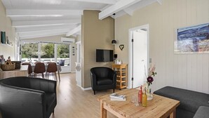 A spacious living area with a dining table, a black leather sofa, and a wooden coffee table. There is a television mounted on the wall and a window providing natural light.