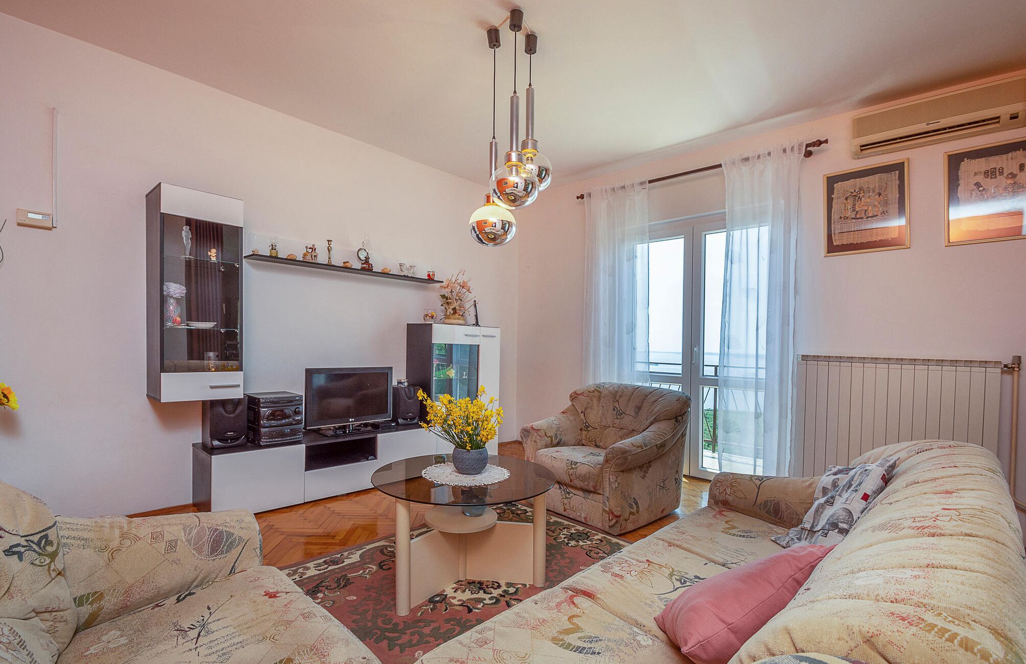 A living room with a sofa, armchair, and a glass coffee table with decorative items. There is a television and a cabinet with decorative items on the wall.