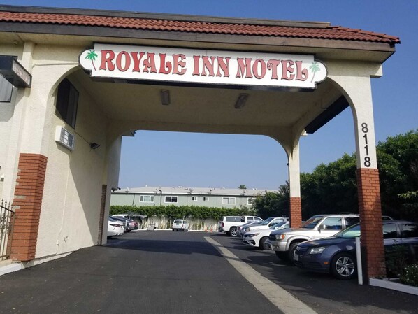 The entrance to the Royale Inn Motel with a clear sign, a parking lot with several cars, and a building in the background. - Royale Inn Motel (Whittier)