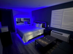 A modern bedroom with a large bed, bedside tables, a small table, and a window with blinds. - Royale Inn Motel (Whittier)