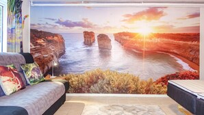 Quarto com um grande mural panorâmico de paisagem costeira, um sofá com almofadas estampadas e um tapete geométrico.