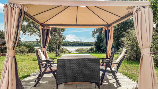 Ein überdachter Außenbereich mit Tisch und Stühlen, umgeben von Vorhängen, mit Blick auf einen See und Grünflächen.