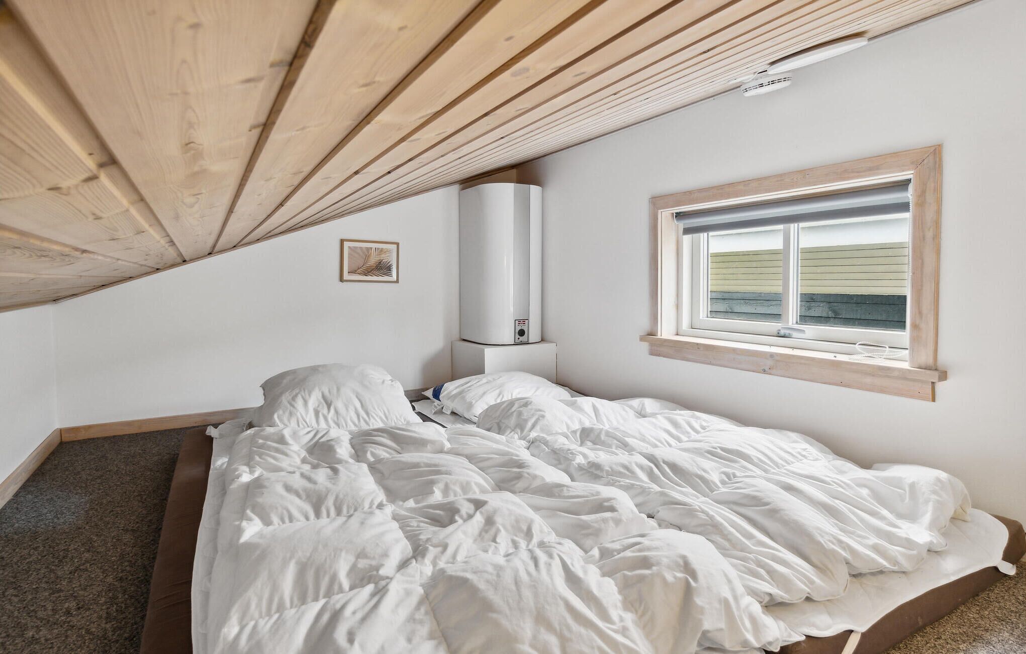 A bedroom with a sloped ceiling, a bed with white bedding, a window with a view, and a wall-mounted heater.