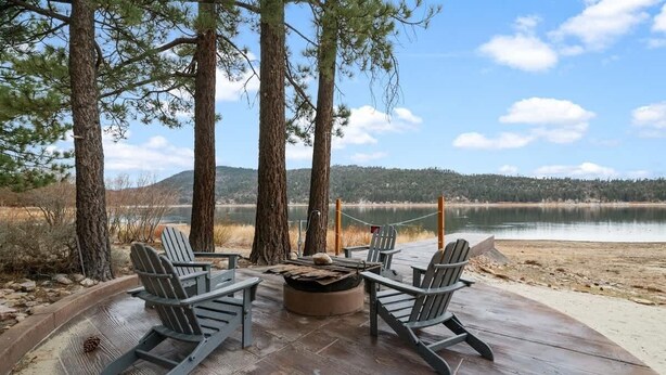 A deck with Adirondack chairs, a fire pit, and a view of a lake and forest. - The Black Cormorant | On the Lake (Big Bear Lake)