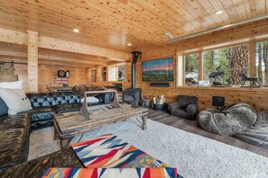 A cozy cabin interior with a wooden ceiling, a fireplace, a sofa, a coffee table, and a large window with a view of trees.